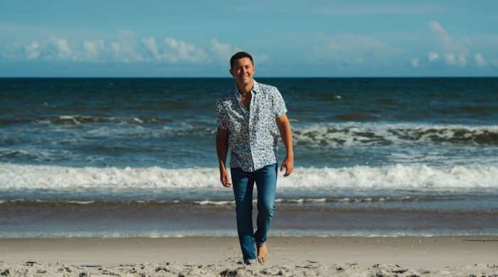 A man walking away from the sea on a beach wearing a blue printed shirt and blue jeans